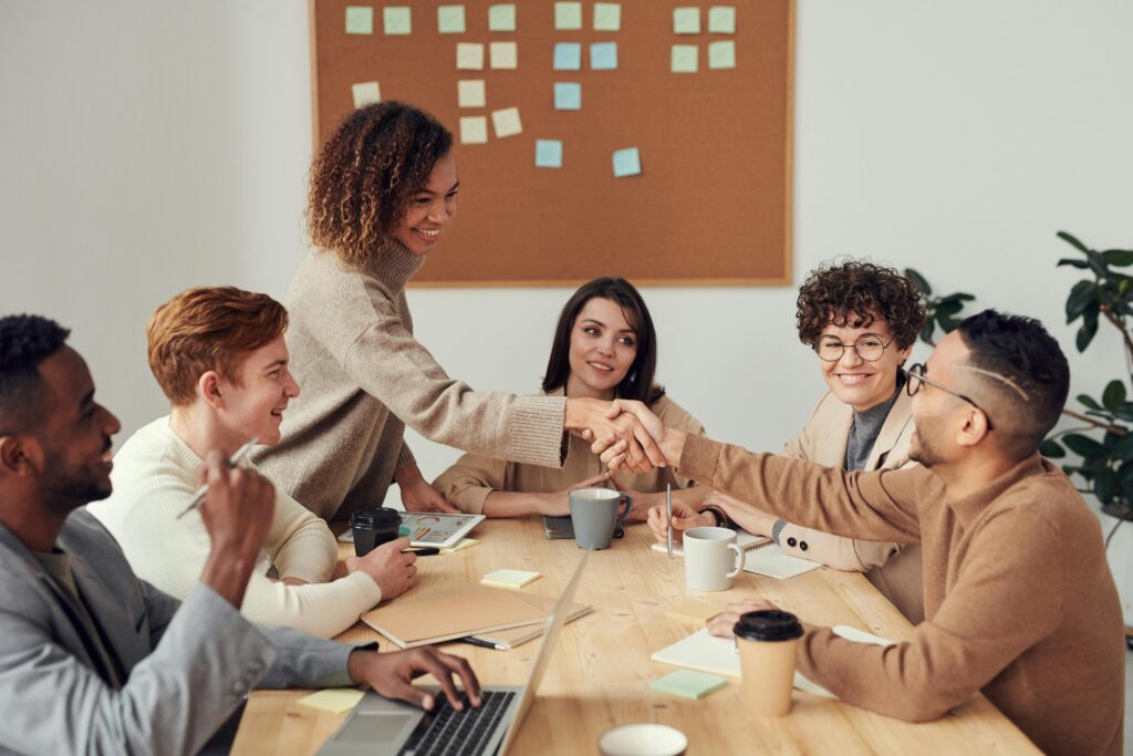 A diverse group of professionals in a business meeting, with two team members shaking hands across a conference table, showing teamwork and collaboration in a modern office setting.